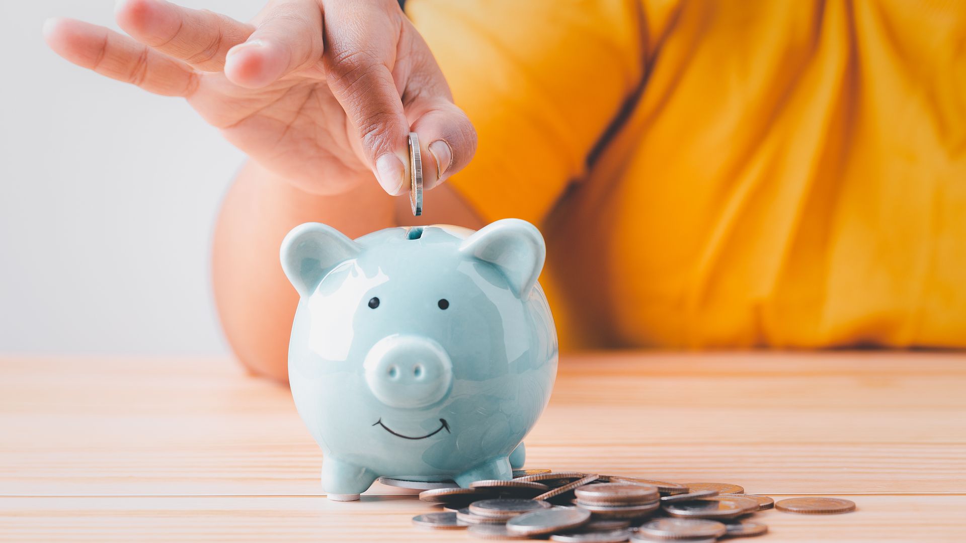 Hand dropping a coin into a light blue piggy bank, with coins scattered on a wooden table